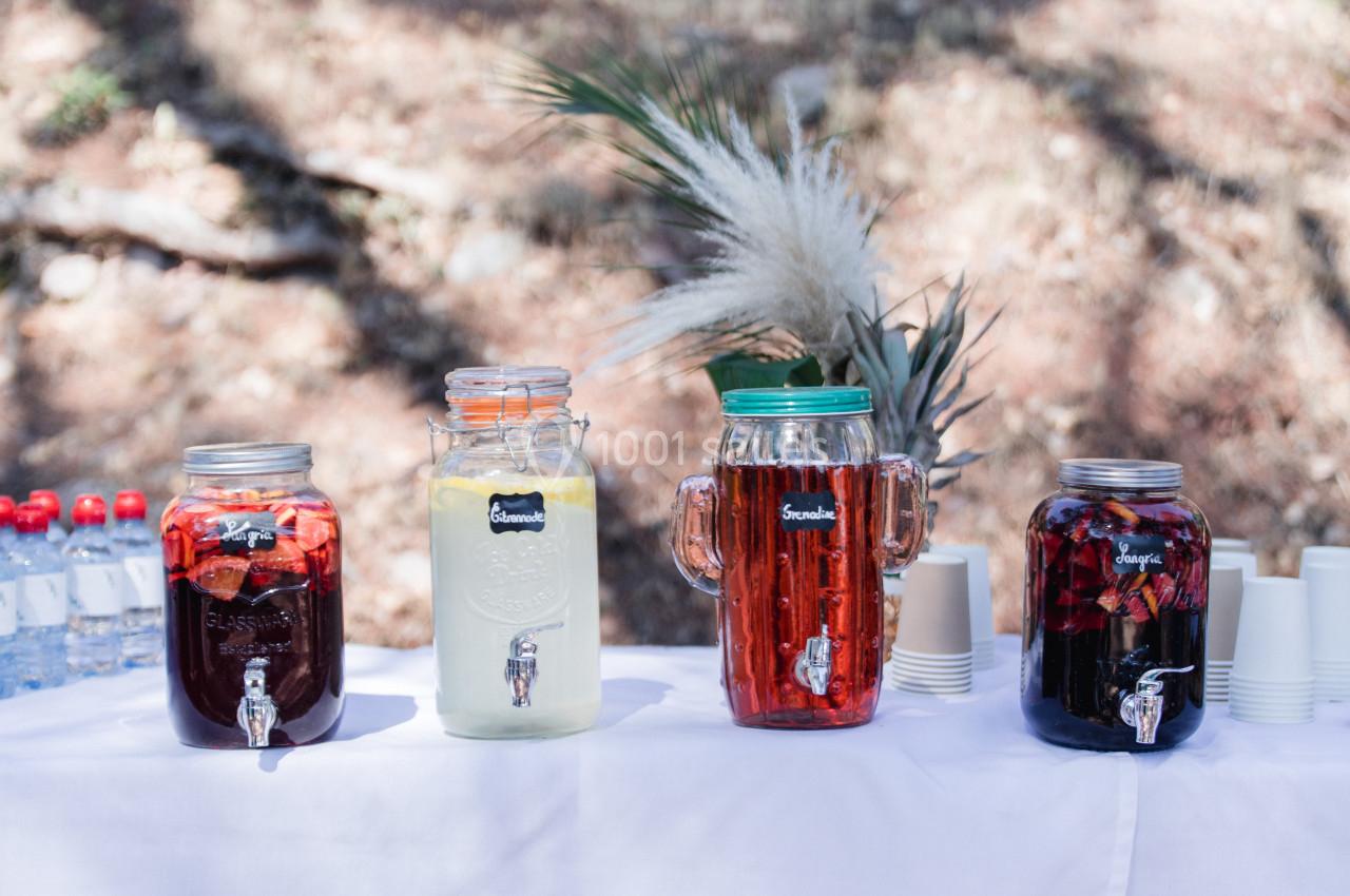 Quatre distributeurs de boissons colorées alignés sur une table blanche en extérieur, avec des gobelets et des bouteilles d…