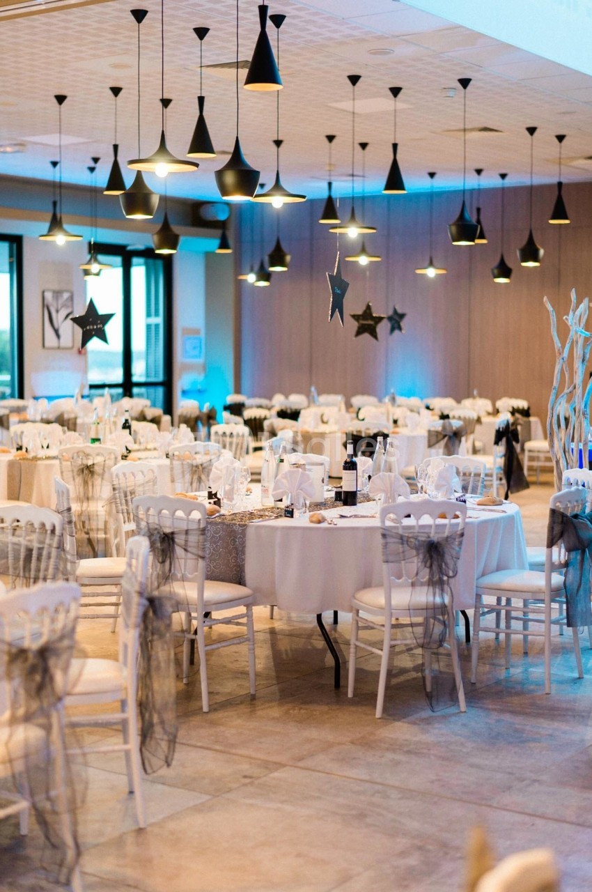 Salle de réception décorée avec des tables rondes dressées, chaises blanches et éclairage suspendu.