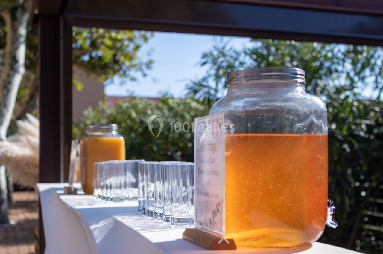 Deux grands bocaux de boissons, l'un orange et l'autre jaune, avec des verres alignés sur une table en extérieur.