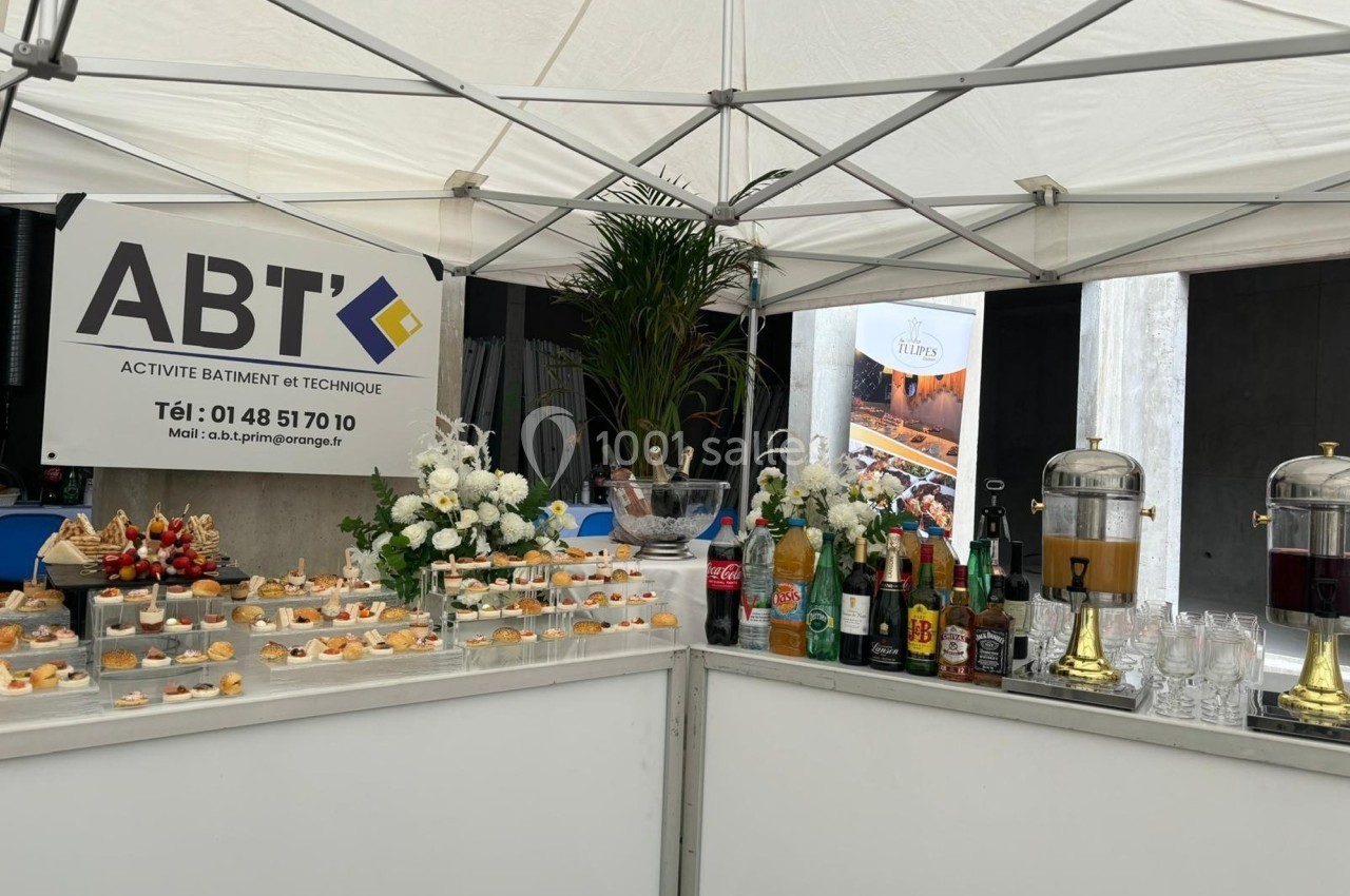 Buffet sous une tente avec amuse-bouches, boissons variées et décoration florale, devant un panneau d'entreprise.