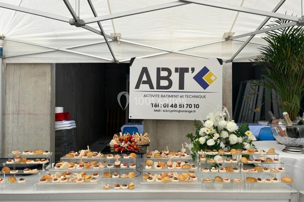Buffet de petits fours et fleurs blanches disposés sous une tente, avec un panneau d'entreprise en arrière-plan.