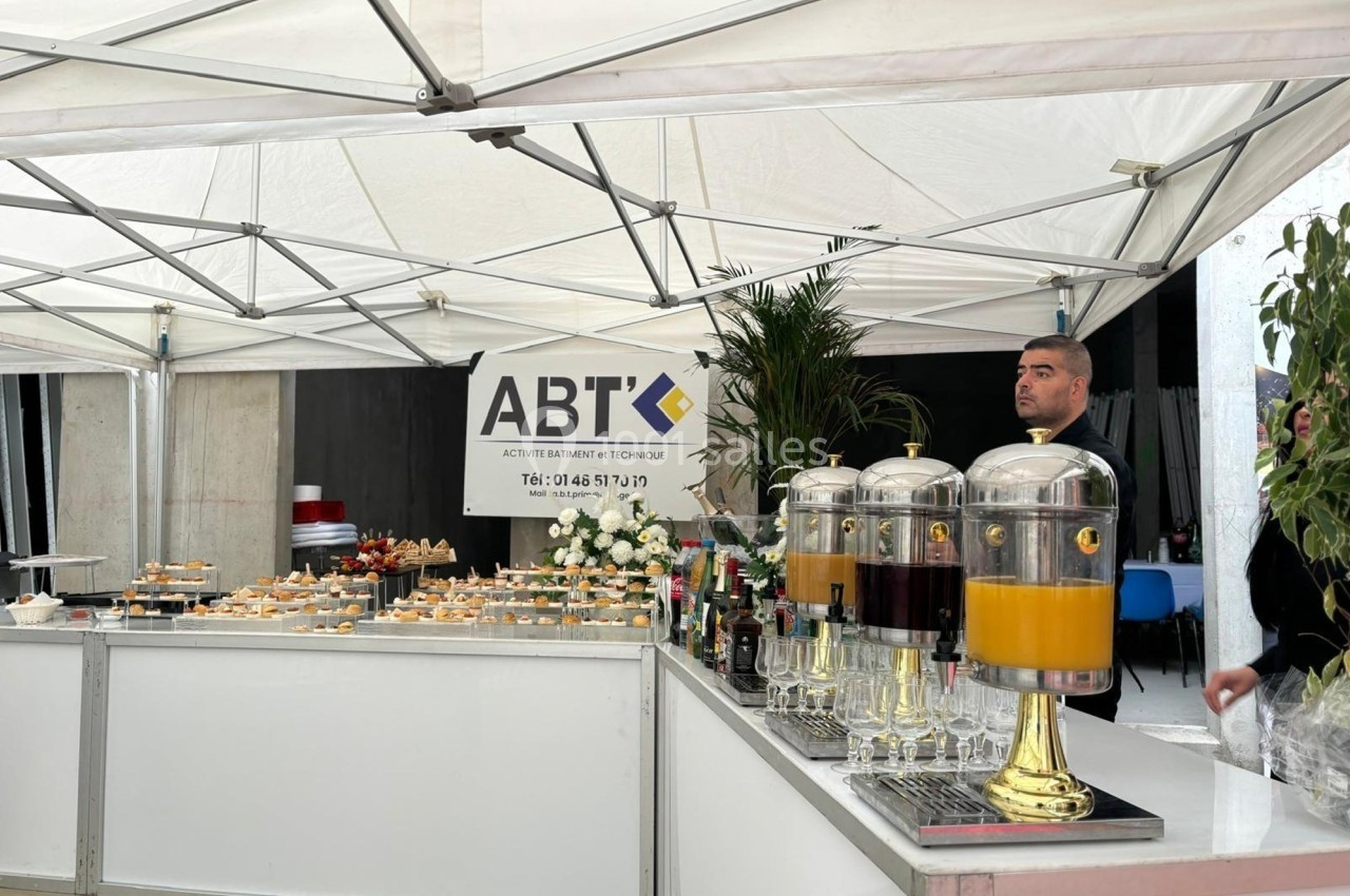 Buffet sous une tente blanche avec boissons en distributeurs, amuse-bouches et un homme derrière le comptoir.