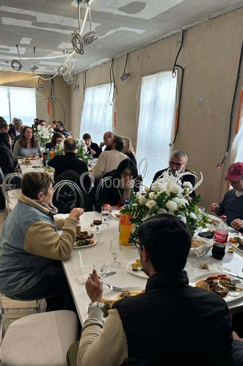 Des personnes partagent un repas convivial autour de longues tables décorées de fleurs dans une salle lumineuse.