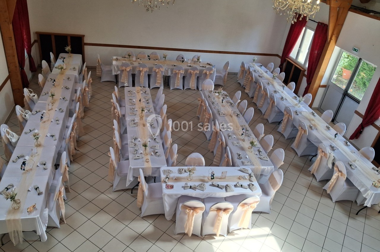 Salle de réception avec tables rectangulaires dressées, nappes blanches et chaises décorées de nœuds beiges.