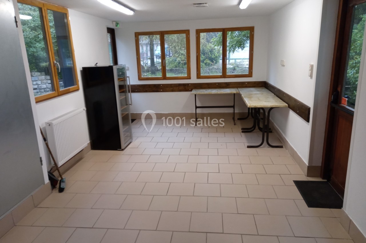 Salle vide avec carrelage beige, murs blancs, fenêtres donnant sur un espace arboré, tables et étagères contre les murs.