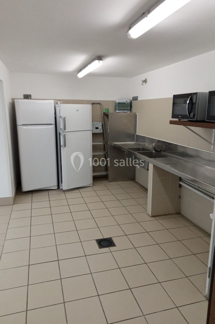 Cuisine professionnelle avec réfrigérateurs, évier en inox, étagères murales et carrelage au sol beige.