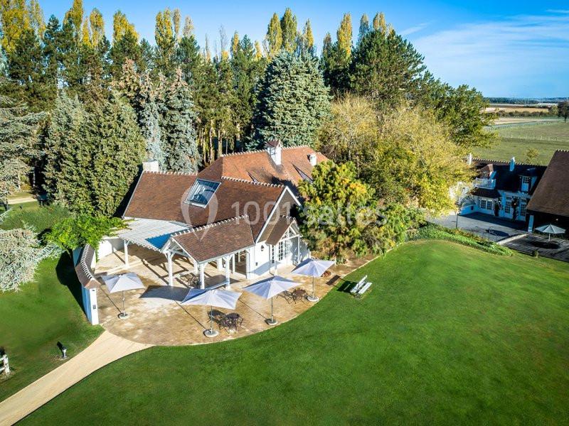 Vue aérienne d'une maison avec terrasse, parasols et jardin, entourée d'arbres et d'un paysage champêtre.