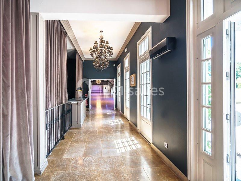 Couloir lumineux avec sol en marbre, murs sombres, lustre suspendu et grandes fenêtres laissant entrer la lumière.