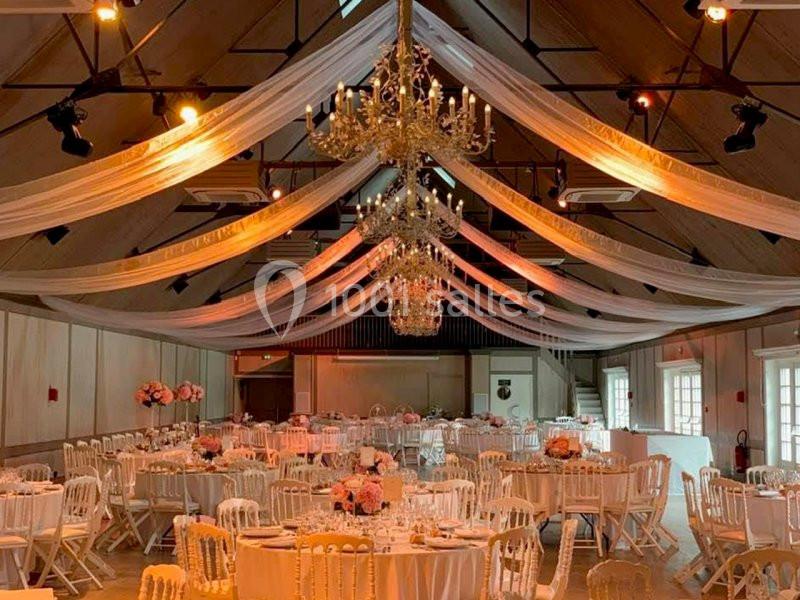 Salle de réception décorée avec des tables rondes, des chaises blanches, des lustres et des drapés suspendus au plafond.