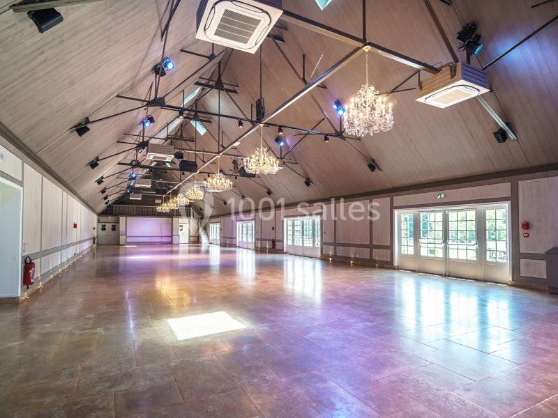 Grande salle lumineuse avec parquet en pierre, lustres suspendus et poutres apparentes sous un plafond en pente.