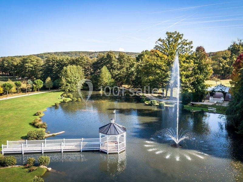 Étang entouré de verdure avec une fontaine centrale et un kiosque accessible par une passerelle en bois.
