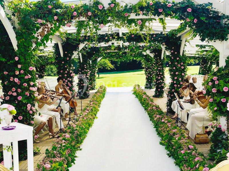 Un quatuor à cordes joue sous une pergola décorée de fleurs roses, bordant une allée blanche dans un jardin.