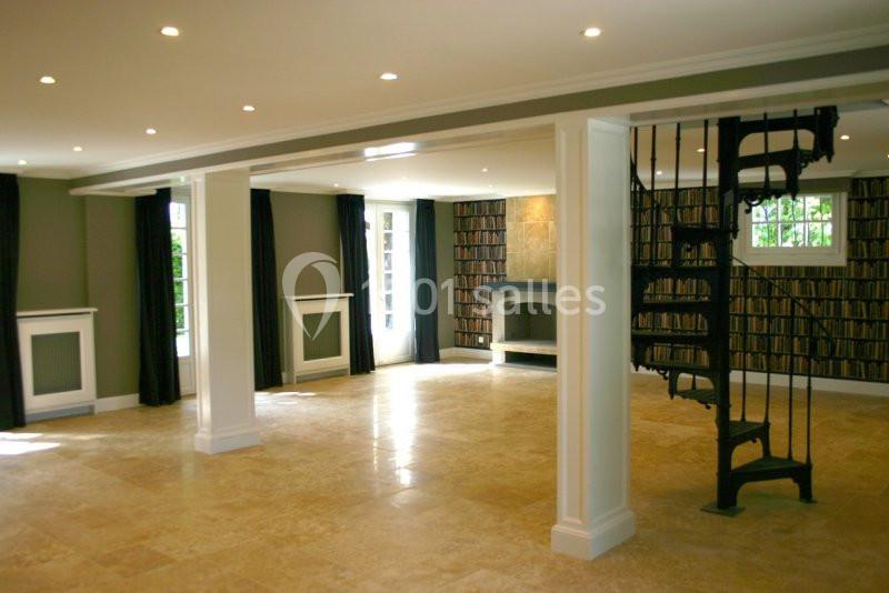 Salle spacieuse avec sol en pierre, escalier en colimaçon, cheminée et murs décorés d'un motif de bibliothèque.