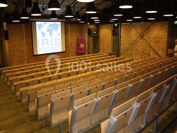 Salle de conférence vide avec des rangées de sièges en bois, un écran de projection et des murs en briques.
