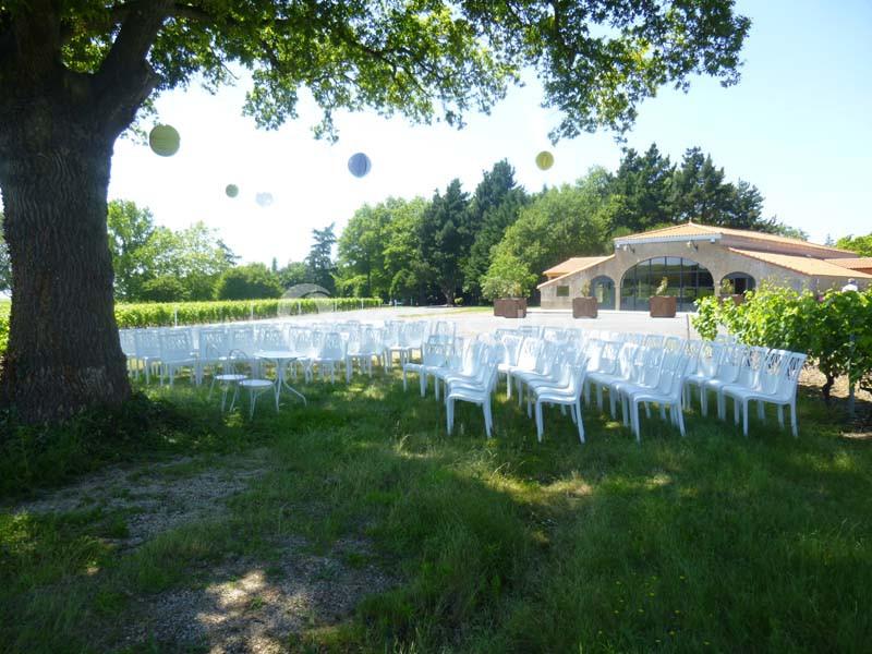 Chaises blanches disposées en extérieur sous un arbre, près d'un bâtiment avec un toit en tuiles, dans un cadre verdoyant.