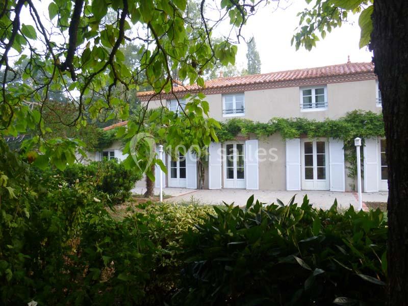 Maison à deux étages avec volets blancs, entourée de végétation dense et d'arbres feuillus.