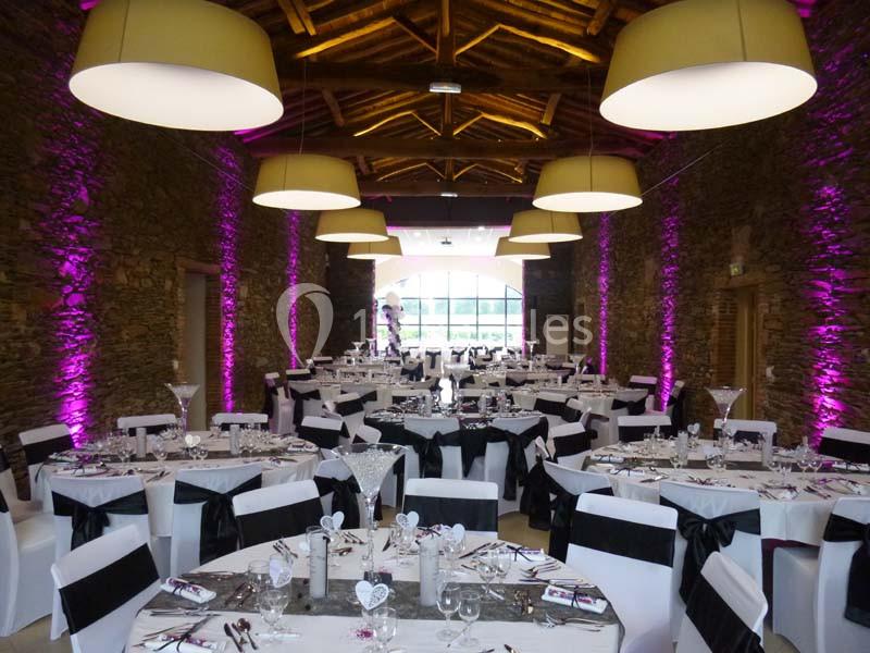 Salle de réception décorée avec des tables rondes, nappes blanches et éclairage violet sur des murs en pierre.