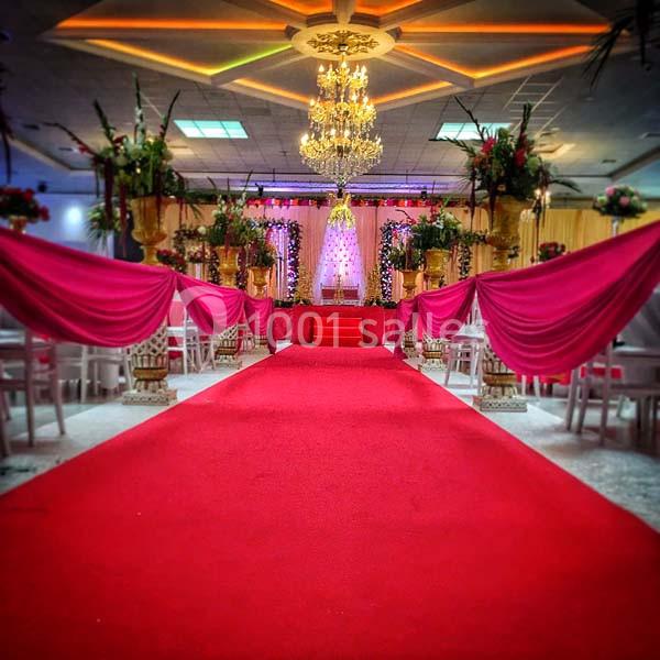 Salle de réception décorée avec un tapis rouge, des drapés roses, des fleurs et un grand lustre central.
