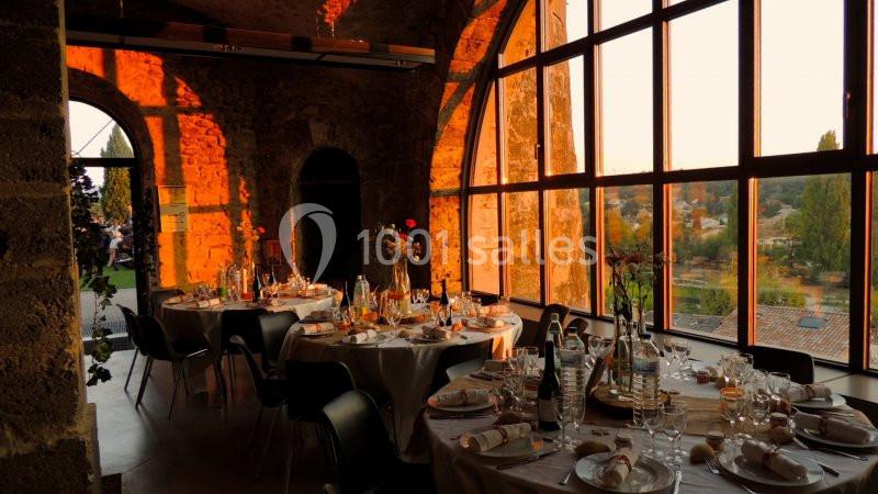 Salle de réception en pierre avec grandes fenêtres, tables dressées et lumière chaude du coucher de soleil.
