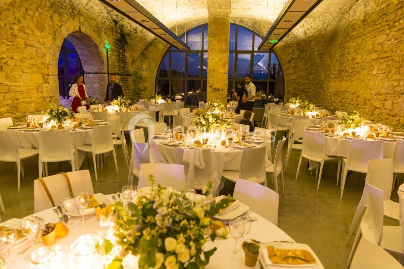 Salle en pierre éclairée, décorée pour un mariage avec des tables rondes, nappes blanches et arrangements floraux.