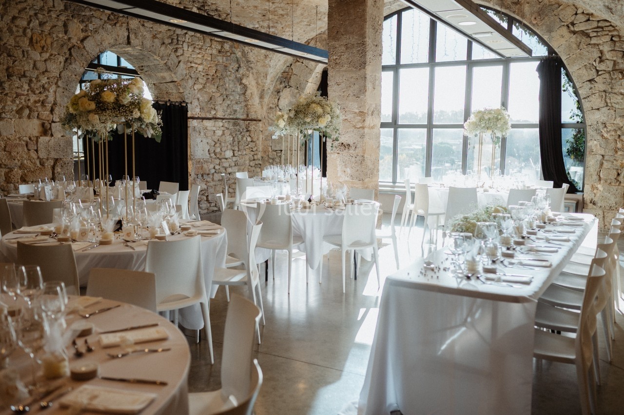 Salle de réception lumineuse avec tables rondes dressées, décorations florales et murs en pierre apparente.