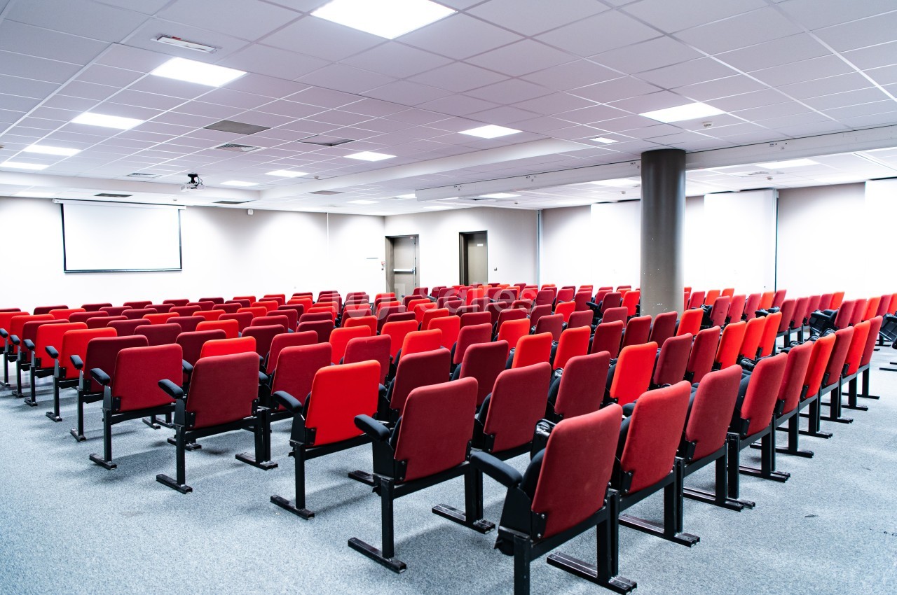 Salle de conférence vide avec rangées de sièges rouges, écran de projection et éclairage fluorescent.