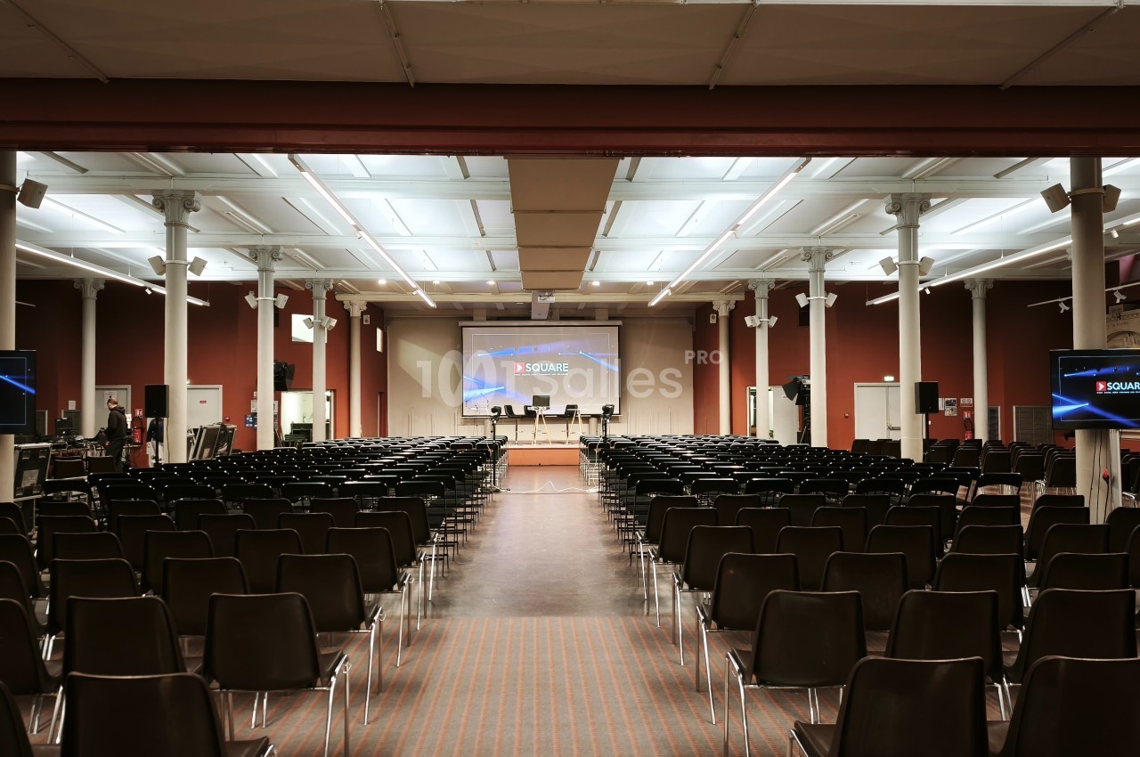 Salle de conférence vide avec des rangées de chaises noires, un écran principal et deux écrans latéraux allumés.