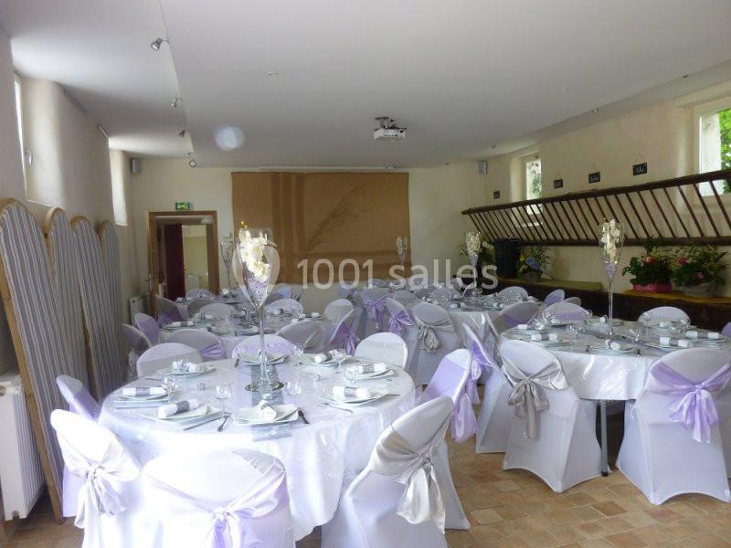 Salle décorée pour un événement avec des tables rondes, nappes blanches, chaises ornées de nœuds mauves et centre de table…