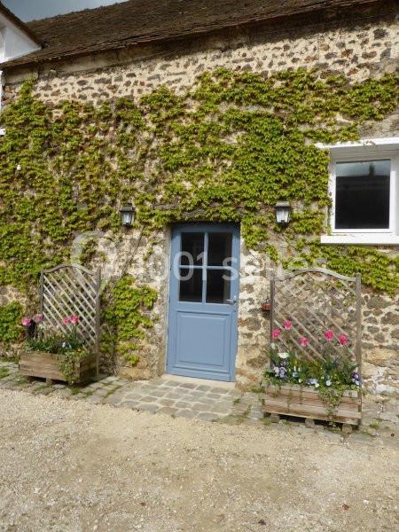 Façade en pierre recouverte de lierre, porte bleue au centre, encadrée de treillis avec des fleurs colorées.