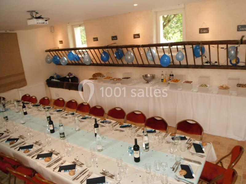 Salle décorée pour un événement avec une longue table dressée, des ballons bleus et un buffet en arrière-plan.