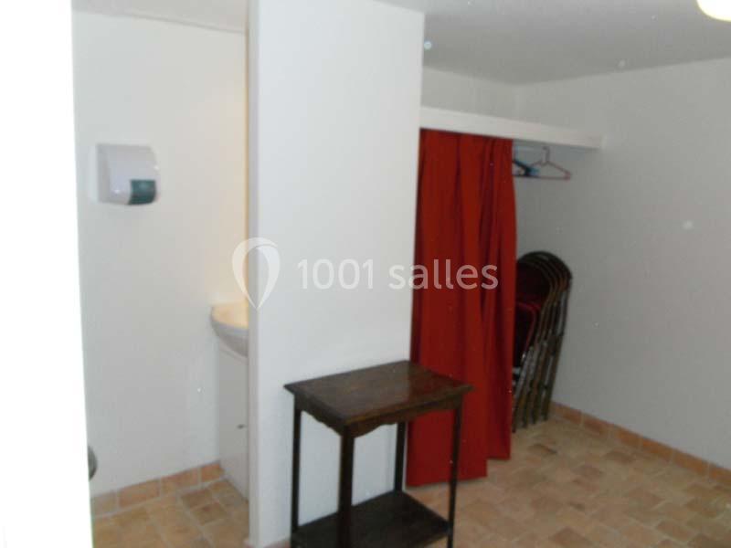 Espace avec un lavabo partiellement caché, rideau rouge, chaises empilées et petite table en bois sur sol carrelé.