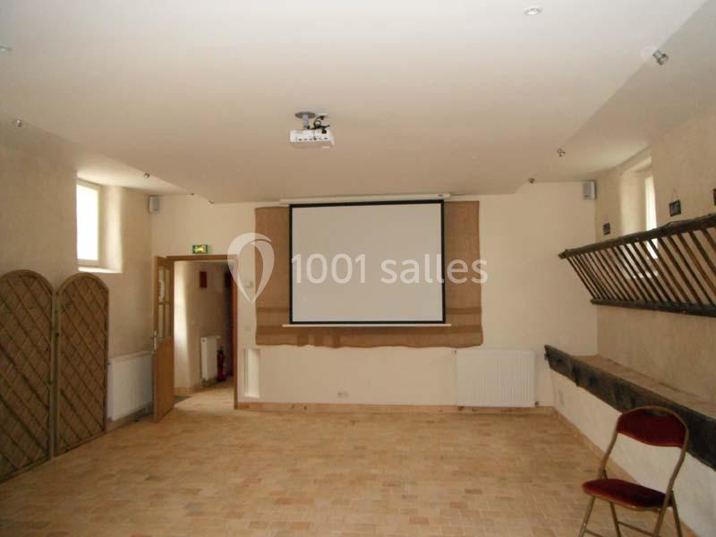 Salle vide avec sol en carrelage, écran de projection, murs clairs, et quelques chaises contre les murs.