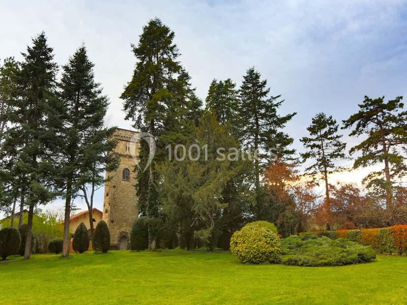 Un jardin verdoyant avec pelouse, arbustes et grands arbres, devant une tour en pierre sous un ciel légèrement nuageux.