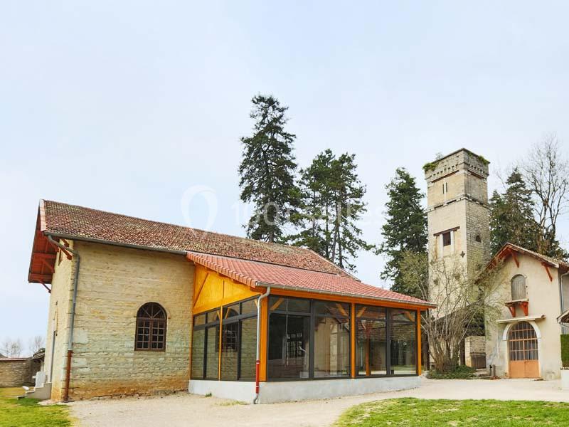 Bâtiment en pierre avec une extension vitrée, entouré d'arbres et d'une tour en arrière-plan.