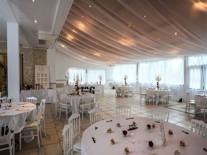 Salle de réception lumineuse avec tables rondes dressées, chaises blanches et plafond décoré de voilages.