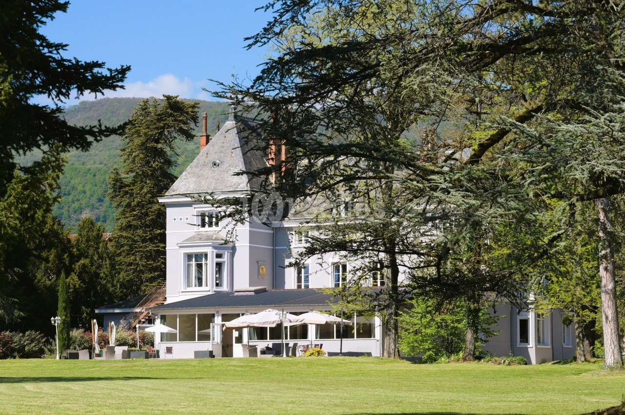 Manoir blanc entouré d'arbres et d'une pelouse, avec terrasse et parasols, au pied de collines verdoyantes.
