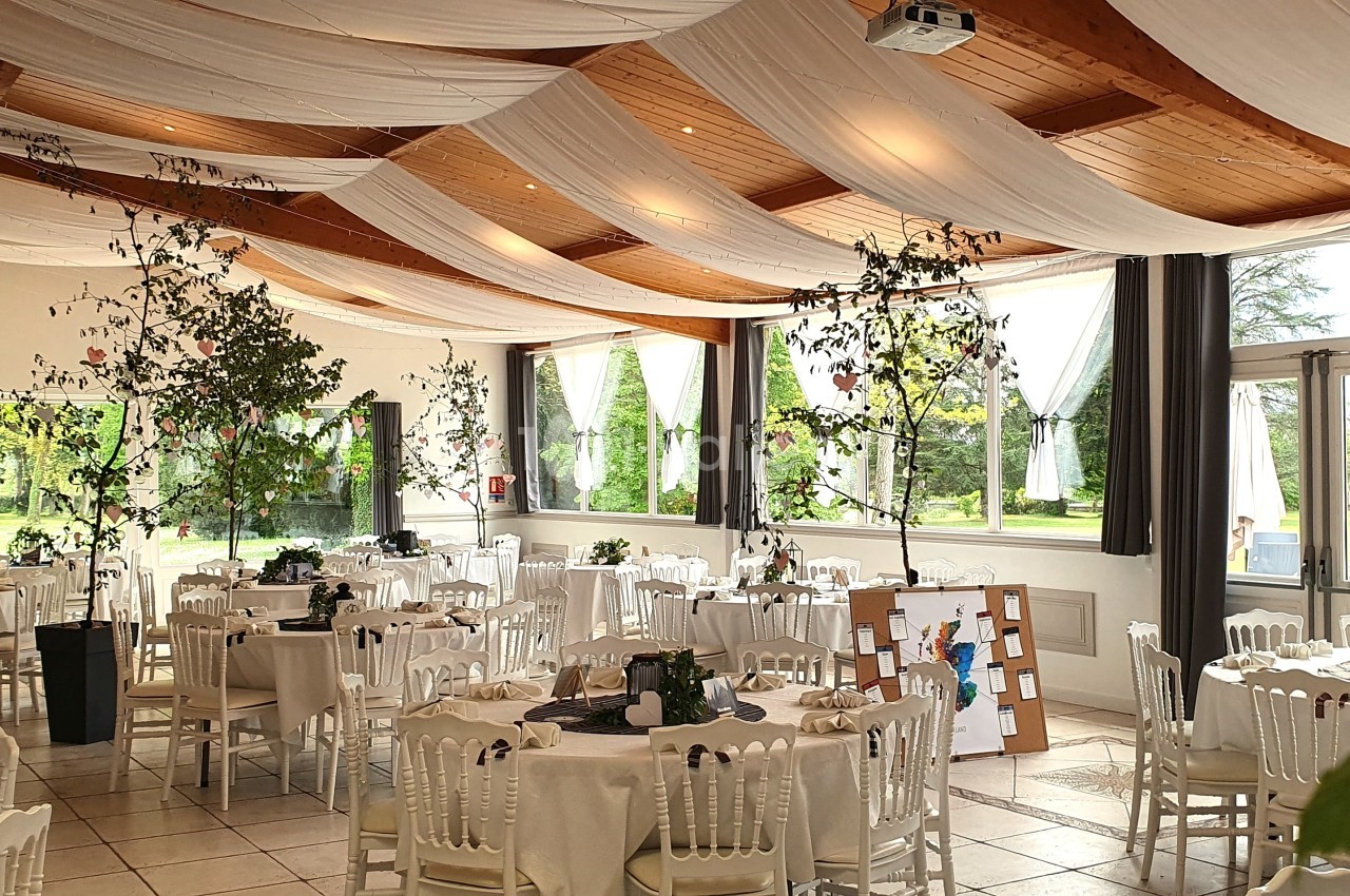 Salle de réception lumineuse avec tables rondes dressées, chaises blanches et décorations végétales suspendues.