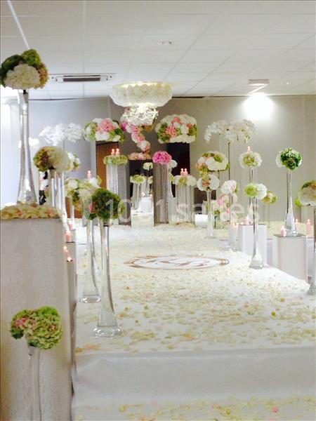 Allée décorée de fleurs colorées sur des vases hauts, avec pétales au sol, dans une salle de réception lumineuse.