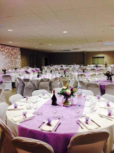Salle de réception décorée avec des nappes violettes, des chaises blanches et des tables dressées pour un événement.