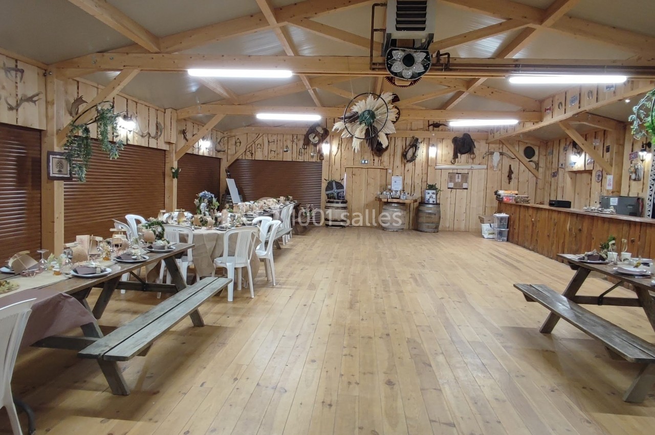 Salle en bois décorée pour un événement, avec tables dressées, bancs, et un bar en fond.