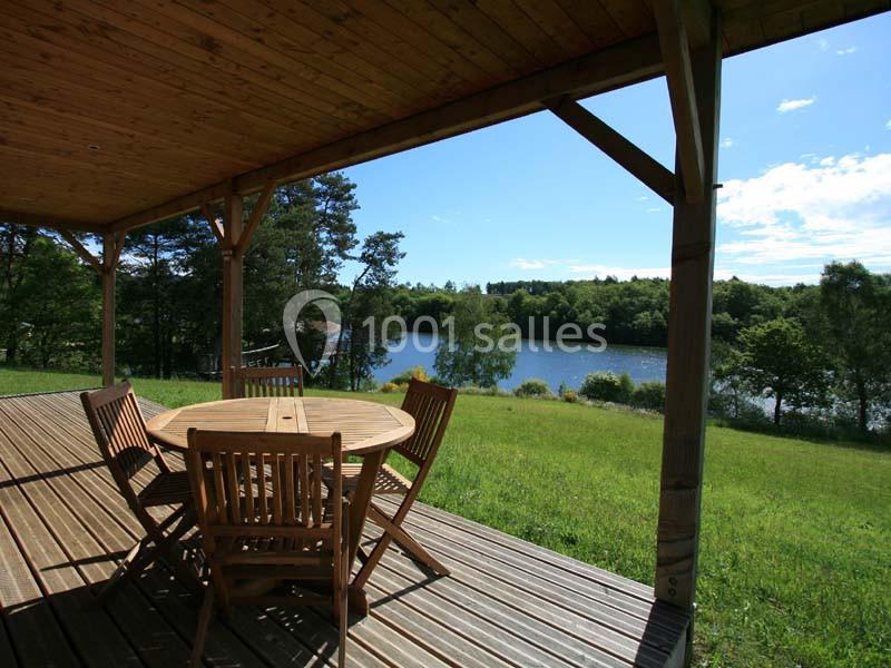 Terrasse en bois couverte avec table et chaises, offrant une vue sur un lac entouré de verdure sous un ciel bleu.