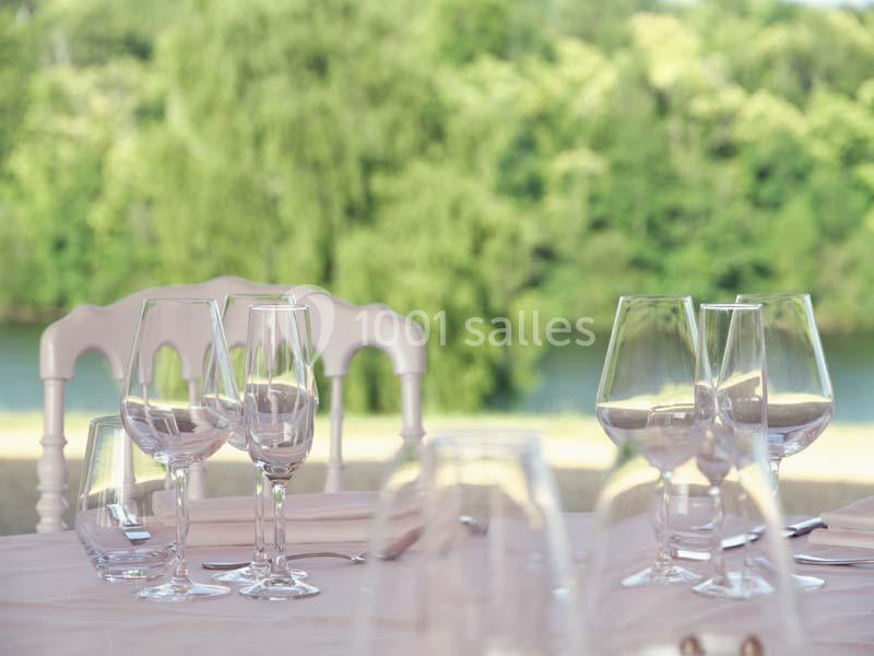 Location salle Meyrignac-l'Église (Corrèze) - Domaine des Monédières #7 Table dressée avec des verres à pied, vue floue sur un paysage verdoyant et une rivière en arrière-plan.
