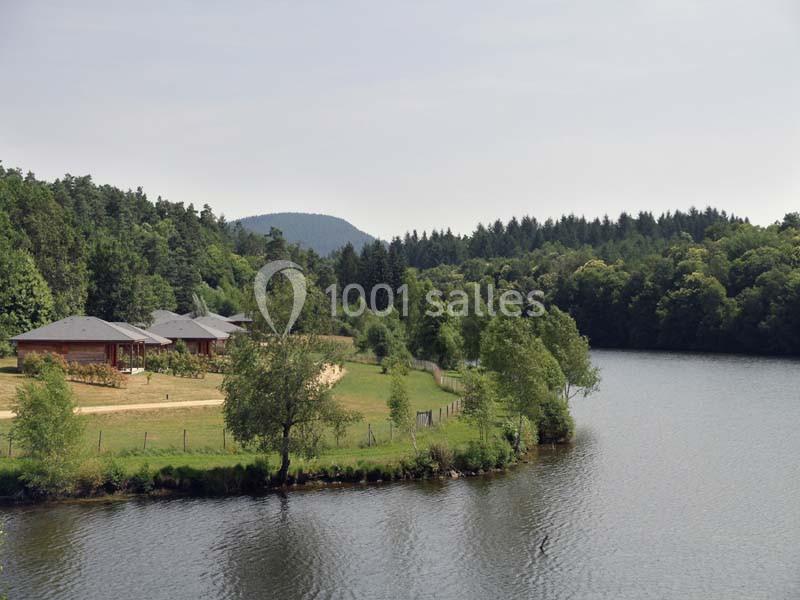 Location salle Meyrignac-l'Église (Corrèze) - Domaine des Monédières #6 Vue d'un lac bordé de forêt avec des chalets en bois et une clairière sur la rive.