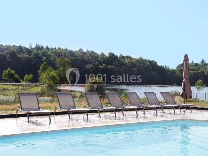 Location salle Meyrignac-l'Église (Corrèze) - Domaine des Monédières #11 Chaises longues alignées au bord d'une piscine avec vue sur un lac et une forêt en arrière-plan.