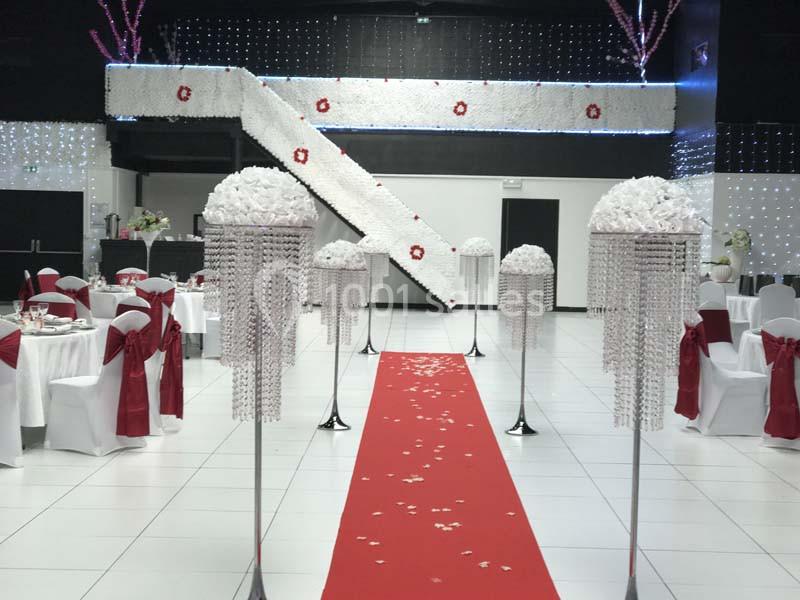 Salle de réception décorée avec des tables dressées, un tapis rouge central et des ornements floraux blancs.