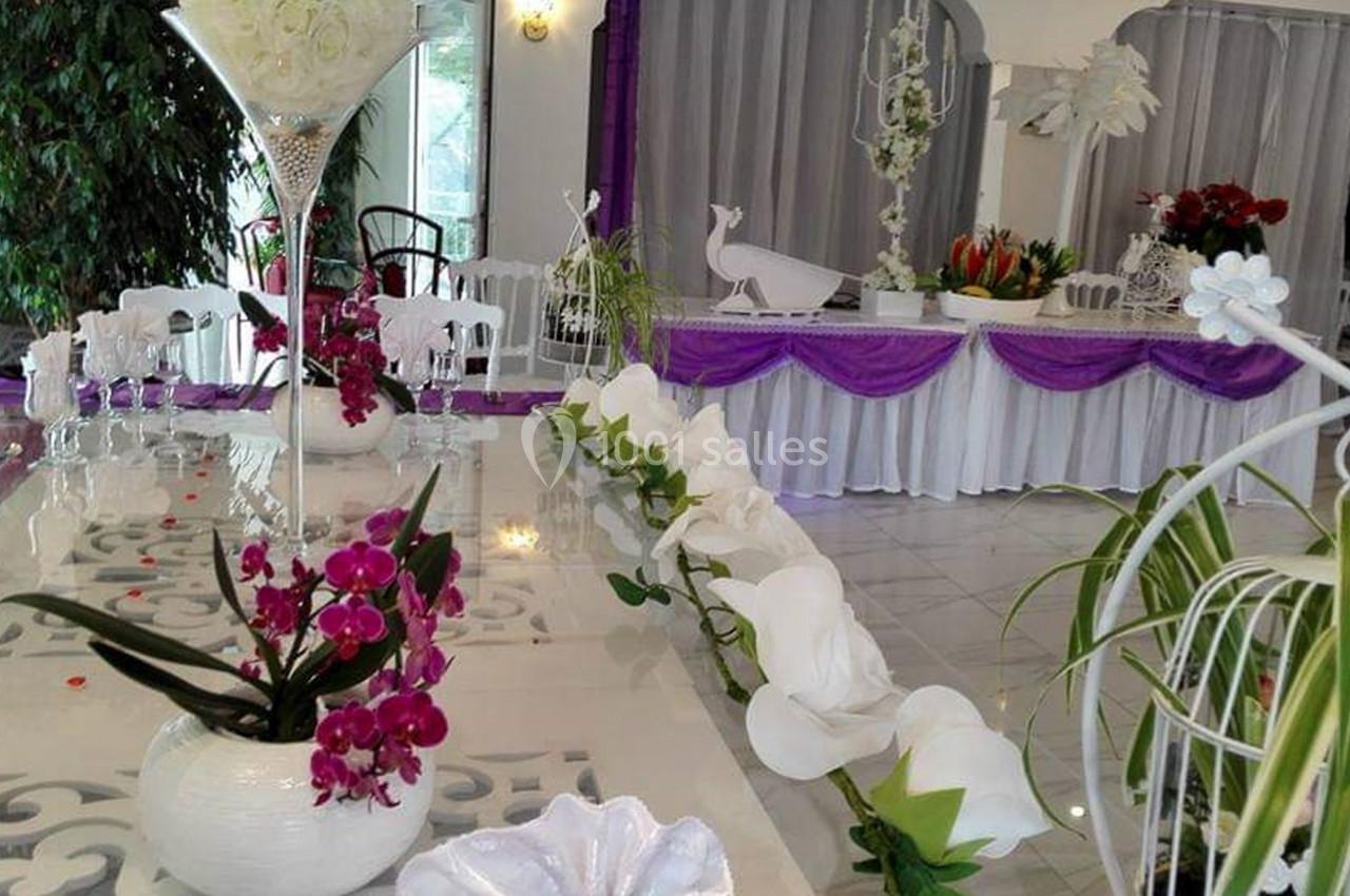 Salle décorée pour un événement avec des fleurs, nappes violettes, et une table ornée de compositions florales.
