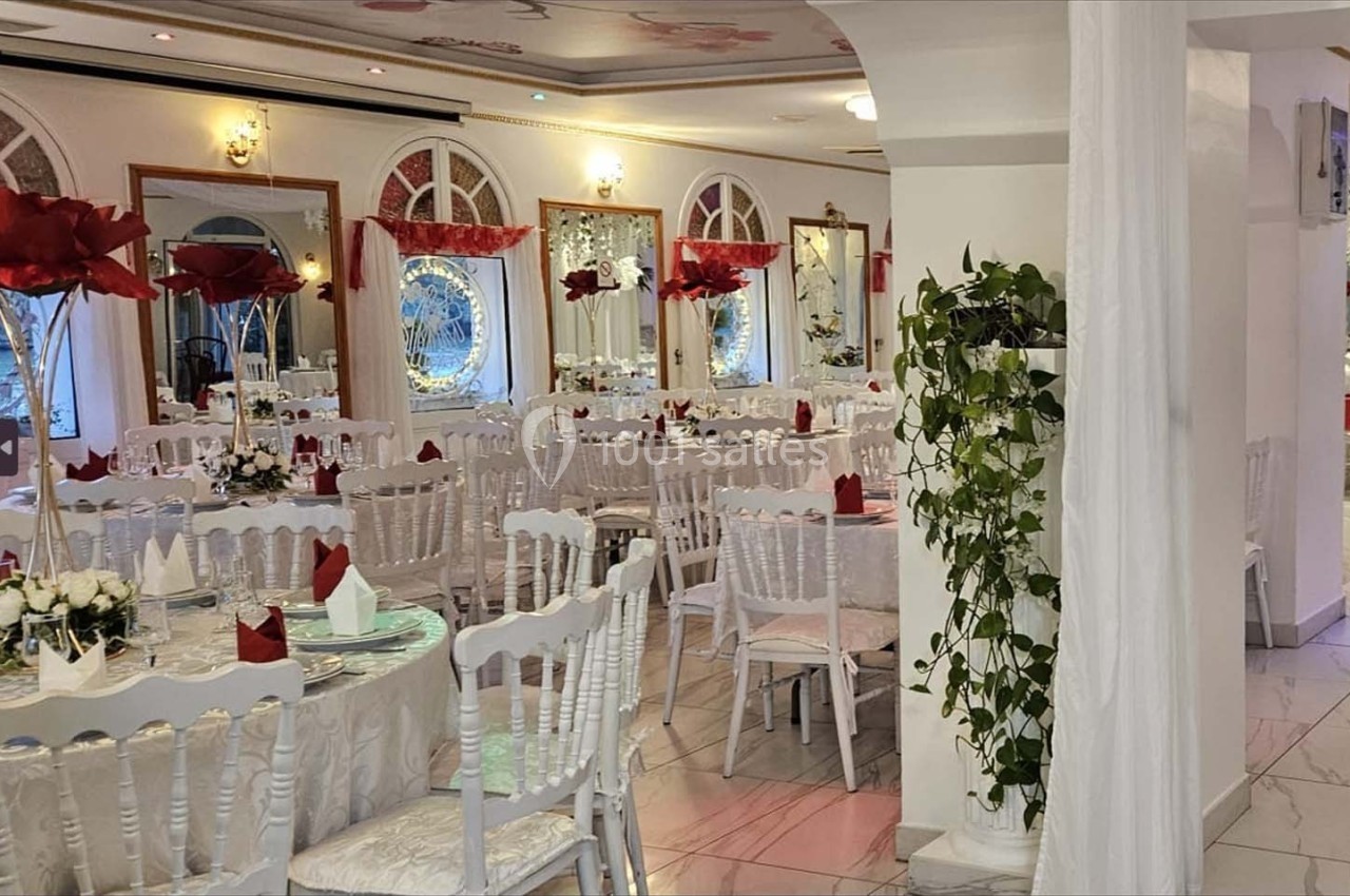 Salle de réception élégante avec tables dressées, chaises blanches, nappes blanches et décorations florales rouges.