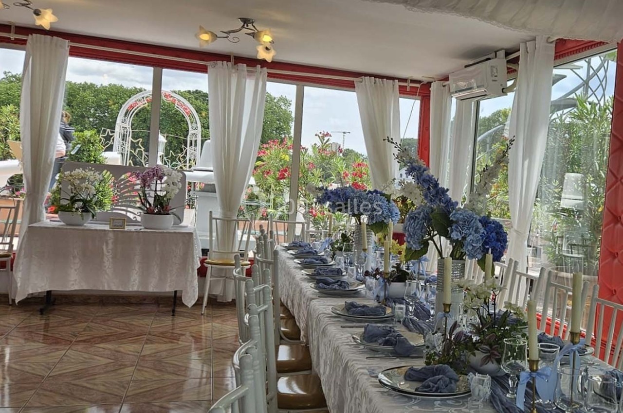 Salle lumineuse décorée pour un événement, avec une grande table ornée de fleurs bleues et blanches près de baies vitrées.
