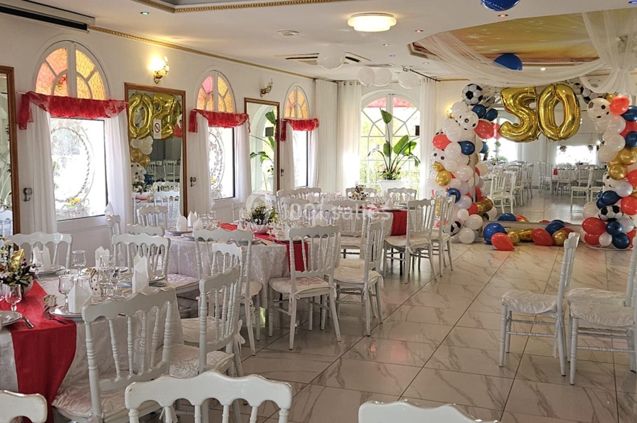 Salle de réception décorée avec des tables dressées, des ballons colorés et des arches pour une fête ou célébration.