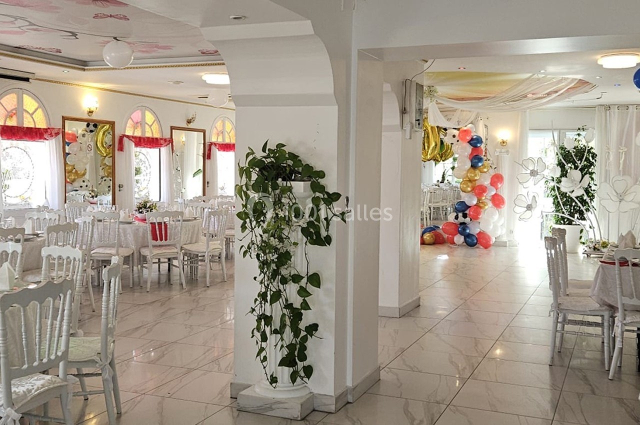 Salle de réception lumineuse décorée avec des tables dressées, des plantes et des ballons colorés au fond.